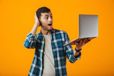 Dizüstü bilgisayar tutan turuncu arka plan üzerinde izole ayakta ekose gömlek giymiş şok genç adam