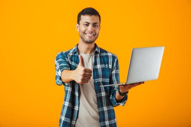 Dizüstü bilgisayar, yaşasın vererek kullanan turuncu arka plan üzerinde izole ayakta ekose gömlek giymiş neşeli genç adam