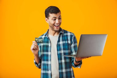 Dizüstü bilgisayarla plastik kredi kartı gösteren turuncu arka plan üzerinde izole ayakta ekose gömlek giymiş neşeli genç adam