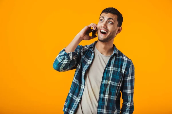 携帯電話で話しているオレンジ色の背景に分離された格子縞のシャツに立って身に着けている陽気な若い男 — ストック写真