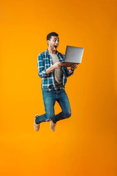 愉快的年轻人穿着格子衬衫跳孤立在橙色的背景 使用笔记本电脑 — 图库照片