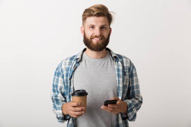 Yakışıklı bir genç sakallı adam cep telefonuyla kahve içme beyaz duvar arka plan üzerinde izole ayakta fotoğrafı.