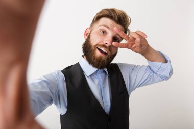 Kamera tarafından barış hareketi gösteren selfie yapmak imge-in bir genç sakallı adam beyaz duvar arka plan üzerinde izole ayakta.
