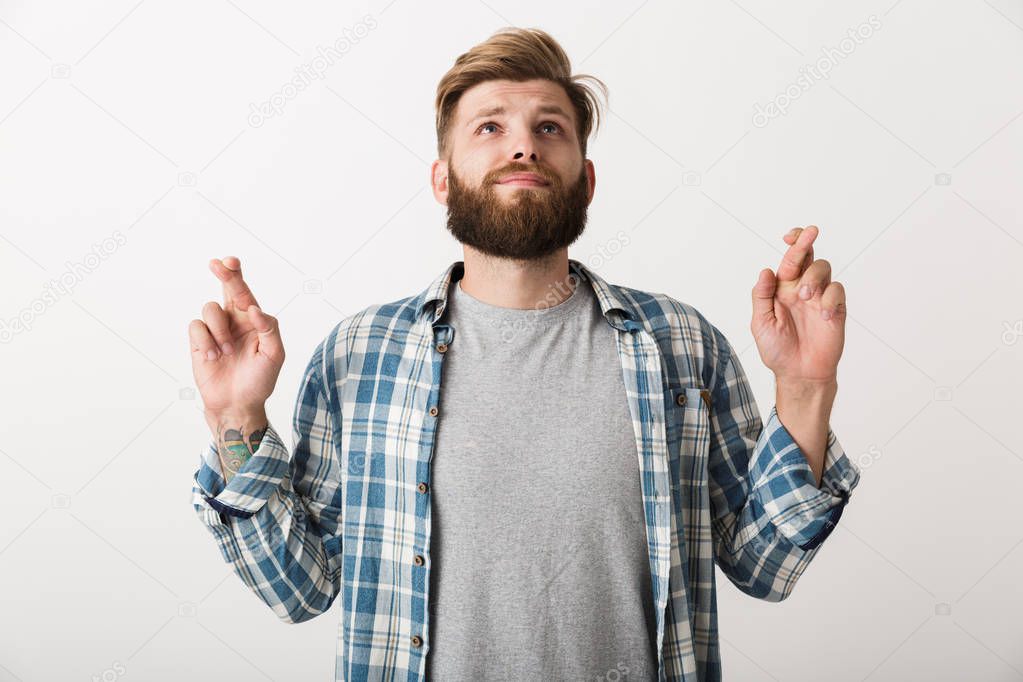 Worried bearded man dressed in plaid shirt standing isolated over white background, holding fingers crossed for good luck