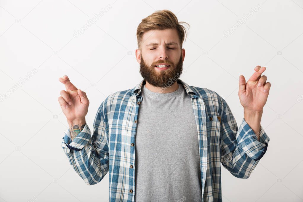 Worried bearded man dressed in plaid shirt standing isolated over white background, holding fingers crossed for good luck