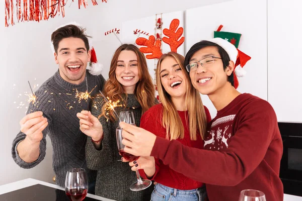 Group of joyful multiethnic people holding glasses with wine and sparklers while celebrating New Year in flat