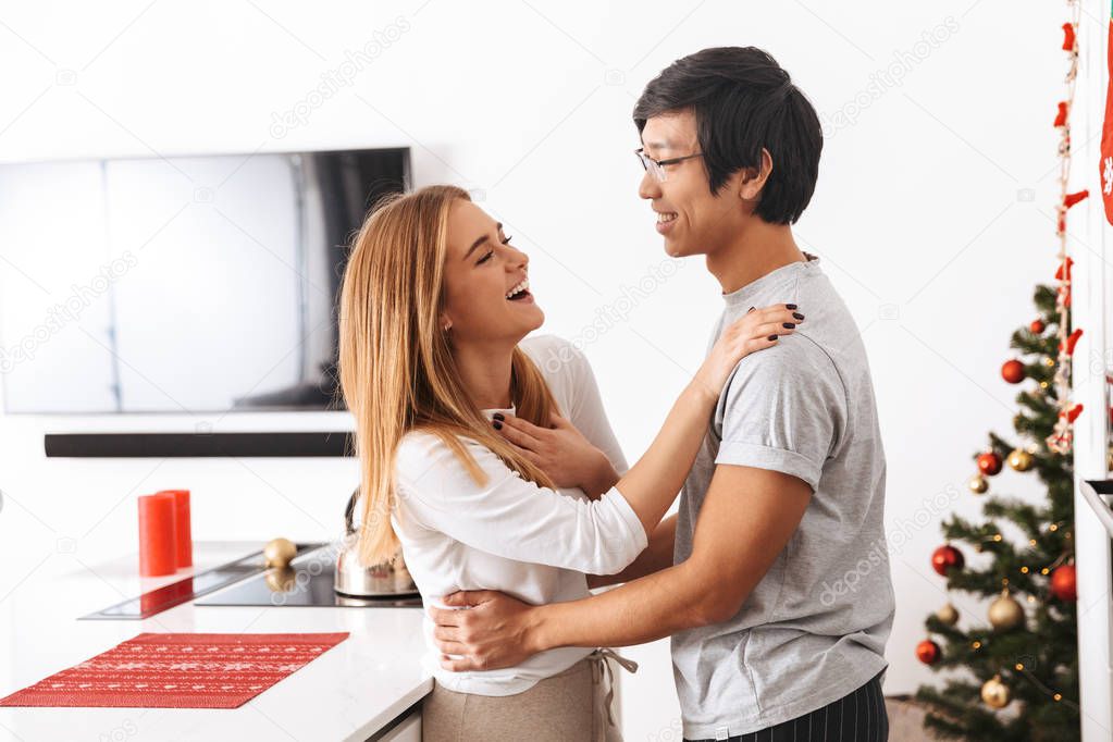 Image of multiethnic couple man and woman standing in bright kitchen and hugging together