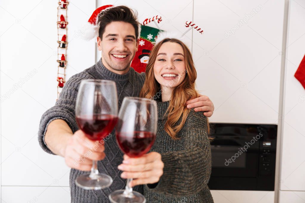 Photo of joyous couple man and woman 20s clinking glasses with wine while celebrating New Year in apartment