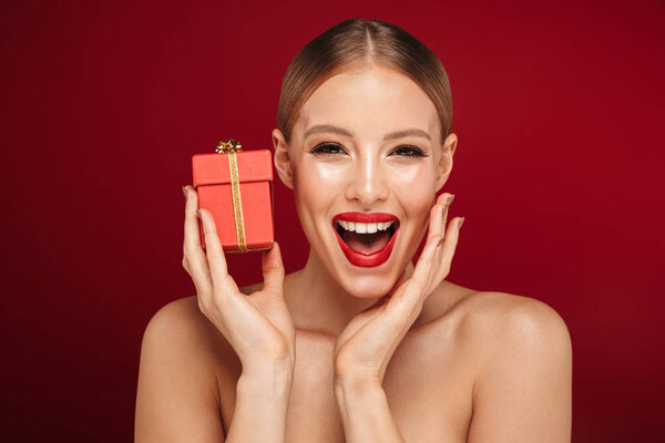 Красота портрет привлекательной блондинки с волосами топлесс женщина стоит изолированно на красном фоне, держа подарочную коробку

