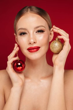 Noel ağacı oyuncaklar tutan izole kırmızı arka plan üzerinde duran bir çekici sarışın saçlı üstsüz kadın güzellik portresi