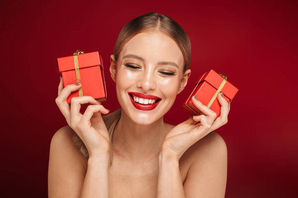 Красота портрет довольно молодой топлесс женщина с макияжем изолированы на красном фоне, показывая две маленькие подарочные коробки
