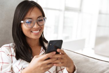 Kanepe rahat daire içinde yatarken modern Asya kadın 20'li yaşlarda holding smartphone fotoğrafı