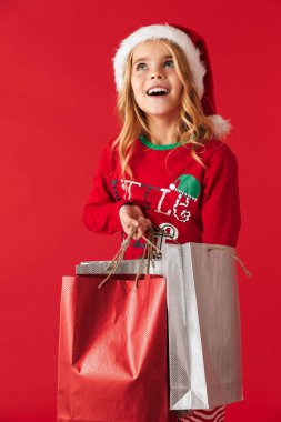 Alışveriş çantaları taşırken Noel şapka ayakta izole kırmızı arka plan üzerinde giyen neşeli küçük kız