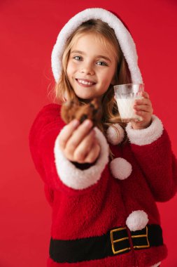 Cam, kurabiye yeme içme sütü izole kırmızı arka plan üzerinde Noel kostüm giyen sevimli Neşeli küçük kız