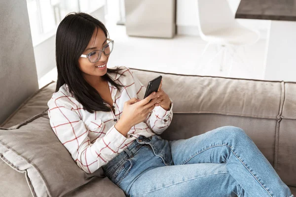Photo of fancy asian woman 20s using cell phone while lying on couch in cozy room