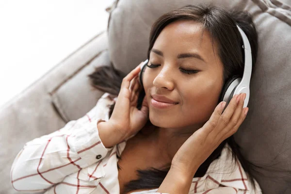 リビング ルームのソファの上に横たわっている間音楽を聴くヘッドフォンをつけた幸せなアジア女の 代の写真 — ストック写真