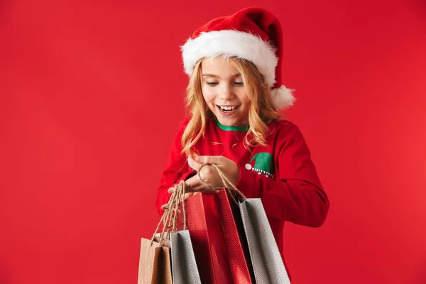 Vrolijk Meisje Kerst Hoed Staande Geïsoleerd Rode Achtergrond Draagt Boodschappentassen — Stockfoto