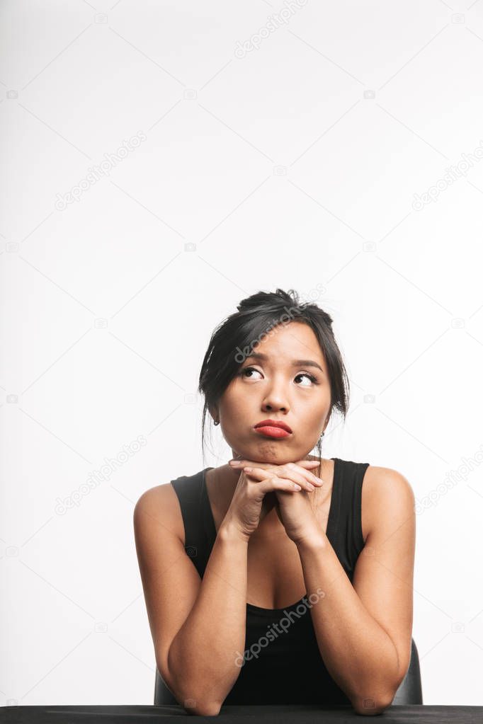 Pensive young asian woman sitting at the table isolated over white background, looking away