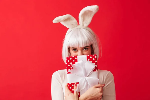 愉快的年轻女孩戴着圣诞兔子耳朵站在孤立的红色背景 拿着礼品盒 — 图库照片