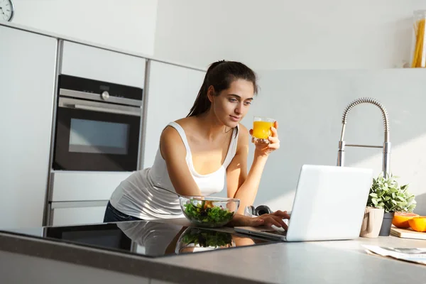 漂亮的年轻女子使用笔记本电脑 而站在厨房和喝橙汁 — 图库照片