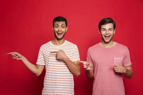 Dois Jovens Alegres Isolados Sobre Fundo Vermelho Apontando Para Espaço — Fotografia de Stock