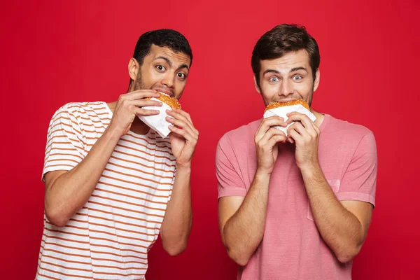 Dos Jóvenes Alegres Pie Aislados Sobre Fondo Rojo Comiendo Hamburguesas —  Fotos de Stock