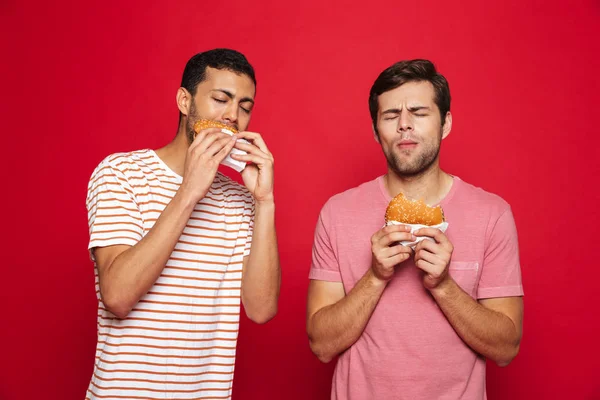 Two delighted men friends standing isolated over red background, eating burgers