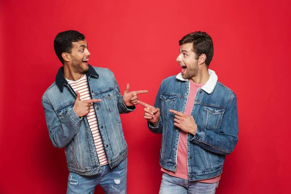 Two excited young men friends wearing denim jackets standing isolated over red background, pointing at each other