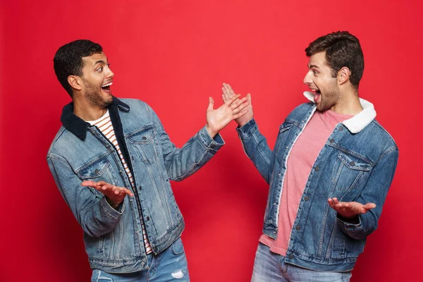 Two excited young men friends wearing denim jackets standing isolated over red background, celebrating