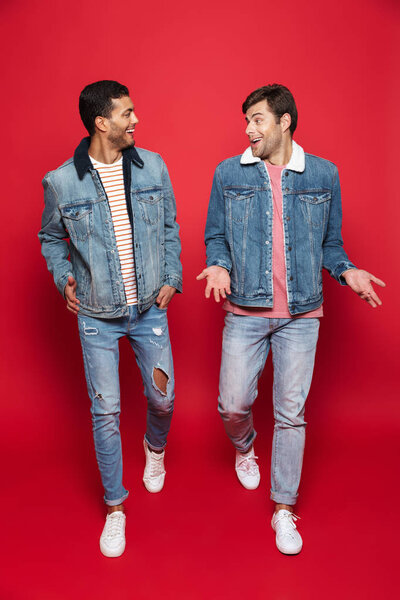 Полная длина двух взволнованных молодых людей друзья носить джинсовые куртки изолированы на красном фоне, ходьба, говорить
