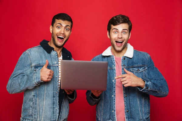 Двоє Веселих Молодих Чоловіків Стоять Ізольовані Червоному Тлі Тримаючи Ноутбук — стокове фото