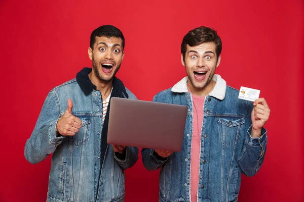 Двоє Веселих Друзів Стоять Ізольовані Червоному Тлі Тримаючи Ноутбук Показуючи — стокове фото