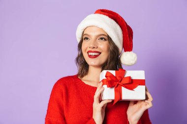 Hediye kutusu tutarak menekşe arka plan üzerinde izole kırmızı Noel şapka ayakta giyen neşeli genç kadın