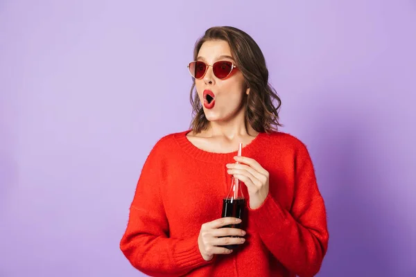 兴奋的年轻女孩穿着红色毛衣和太阳镜站在孤立的紫罗兰色背景 喝从玻璃瓶用吸管 — 图库照片