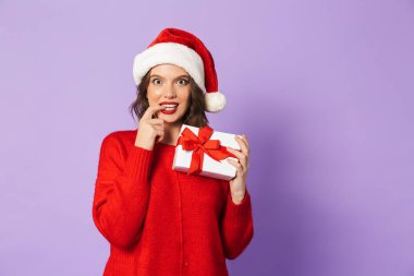 Hediye kutusu tutarak menekşe arka plan üzerinde kırmızı Noel şapka ayakta giyen gülümseyen dalgın genç kadın izole