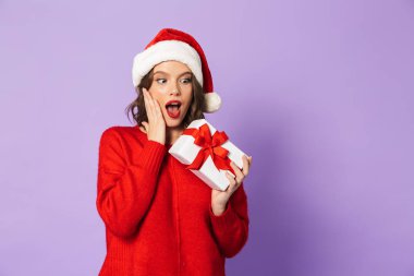 Hediye kutusu tutarak menekşe arka plan üzerinde izole kırmızı Noel şapka ayakta giyen neşeli genç kadın