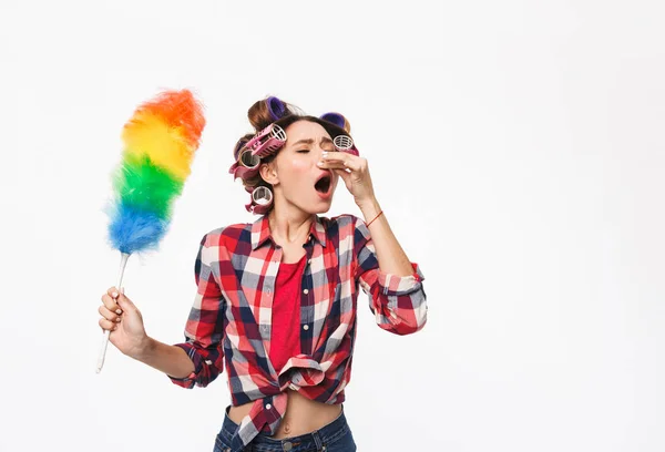 Busy housewife with curlers in hair standing isolated over white background, holding duster, unpleasant smell