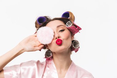 Toz puf kullanarak güzel ev kadını curlers bornoz ayakta beyaz arka plan üzerinde izole giyen saç ile