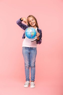 Dünya küre tutan pembe arka plan üzerinde izole bir gülümseyen küçük kız ayakta tam uzunlukta