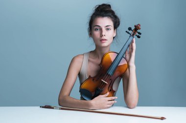 Mavi arka plan üzerinde izole bir keman ile masada oturan güzel bir genç kadının portresi