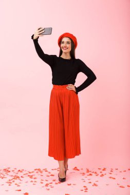 Tam uzunlukta bir selfie alarak pembe arka plan üzerinde izole kırmızı bere ayakta giyen güzel genç bir kadın portresi