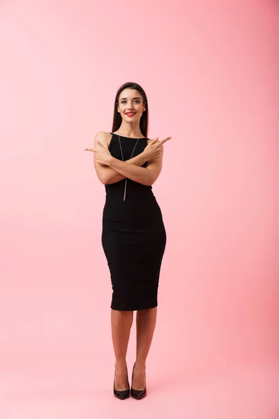 一个美丽的年轻女子穿着黑色礼服站在粉红色的背景隔离 指着复制空间的全长肖像 — 图库照片