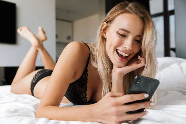 Cep Telefonu Kullaranarak Yatakta Döşeme Çamaşırı Giyen Güzel Gülümseyen Genç — Stok fotoğraf