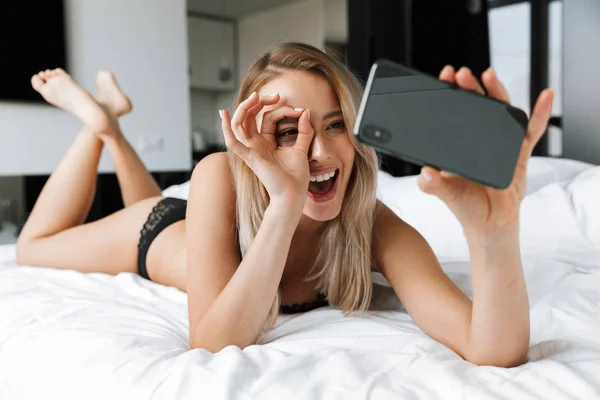 Güzel genç kadın iç çamaşırı iç giyim sabah yatakta evde cep telefonuyla bir selfie almak. — Stok fotoğraf