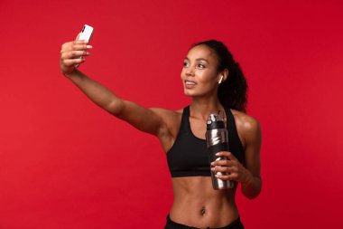 Görüntü izole kulaklık ile müzik dinleme kırmızı duvar arka plan üzerinde bir güzel genç Afrika spor fitness kadının poz tarafından cep telefonu ile su şişe tutan bir selfie almak.