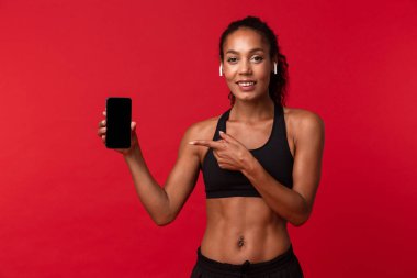 Akıllı telefon ve earpods kırmızı arka plan üzerinde izole kullanarak siyah spor giyim güzel Afrikalı-Amerikalı kadın portresi