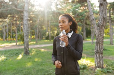 Çekici kadın 20'li yaşlarda giyen siyah eşofman ve kulaklık yeşil park boyunca yürürken İçmesuyu görüntüsünü