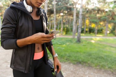 Atletik kadın 20'li yaşlarda giyen siyah eşofman ve kulaklık yeşil park smartphone kullanarak kırpılmış görüntü