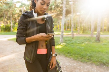 Çekici kadın 20'li yaşlarda giyen siyah eşofman ve kulaklık yeşil park smartphone kullanarak görüntü