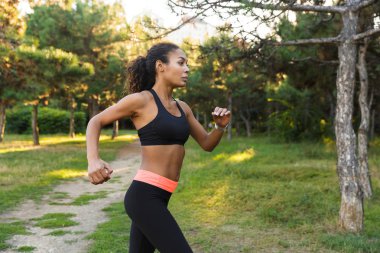 Beautiful african american woman 20s wearing black sportswear running through green park clipart